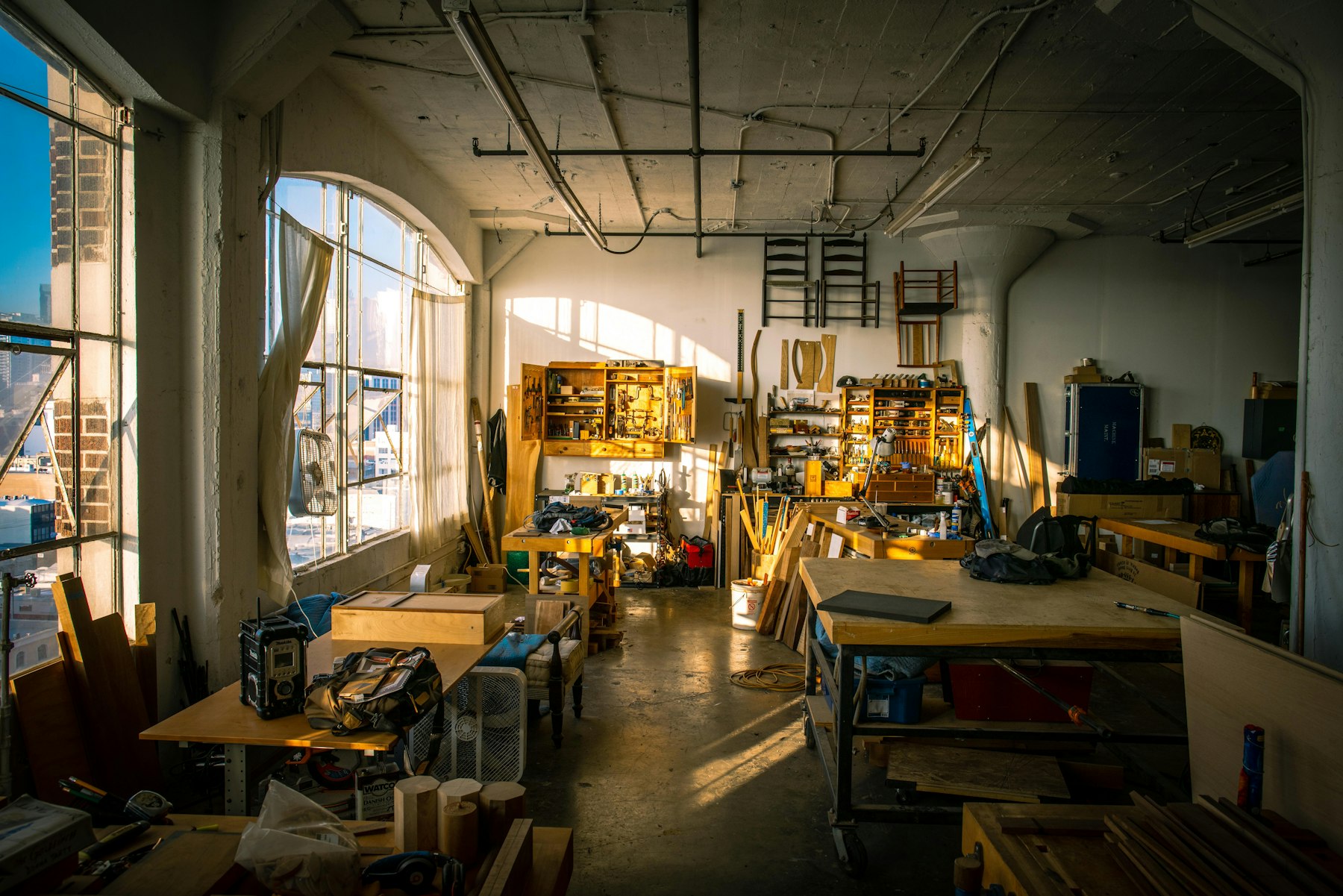A sunlit workshop with long wooden workbenches, tools and tall windows